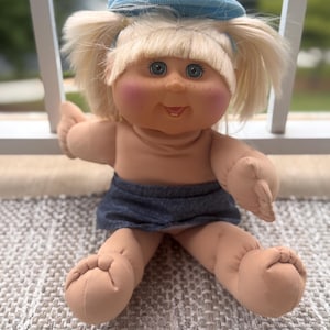 May include: A soft-sculpture doll with blonde yarn hair styled in pigtails, wearing a light blue fabric cap and a dark blue denim-style skirt. The doll features blue eyes, rosy cheeks, and freckles, with a light skin tone and a small open mouth.