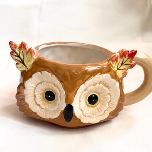 May include: A ceramic mug shaped like an owl head with a glossy brown finish, large cream-colored eyes, a black beak, and two decorative autumn leaves in red, orange, and yellow attached to the top rim, featuring a smooth beige handle.