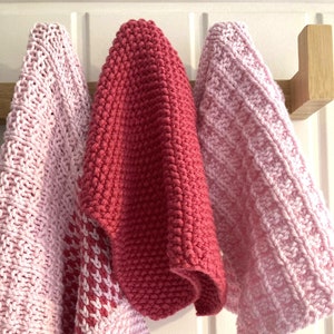 Op de afbeelding: Drie gebreide theedoeken hangen aan een houten rek. Eén is roze, één is lichtroze met een wit en roze patroon en één is felrood.