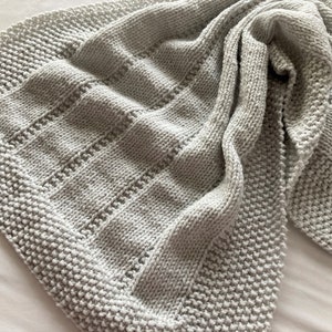 May include: A light grey knitted blanket featuring a textured border of seed stitch and a central panel of alternating vertical stockinette stitch columns separated by narrow rows of eyelet lace detailing, creating a soft and uniform appearance.