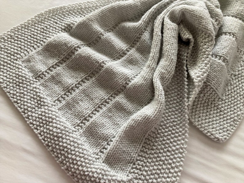 May include: A light grey knitted blanket featuring a textured border of seed stitch and a central panel of alternating vertical stockinette stitch columns separated by narrow rows of eyelet lace detailing, creating a soft and uniform appearance.