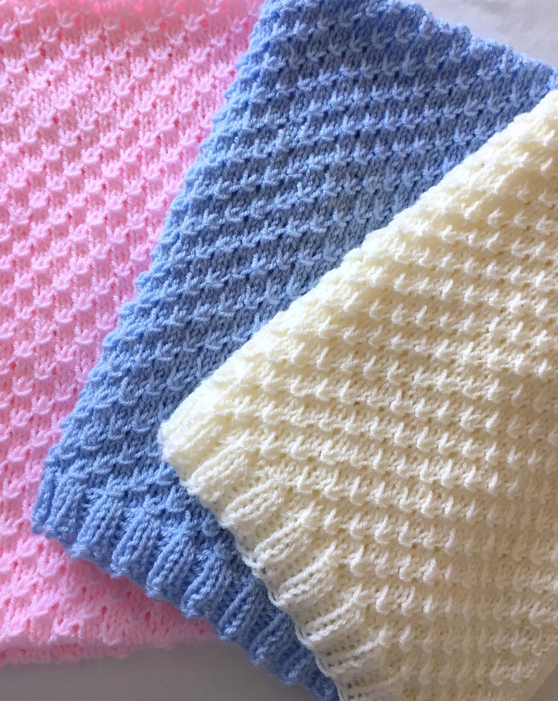 May include: Three knitted baby blankets in pink, blue, and white. The blankets are made with a textured stitch and are folded on top of each other.