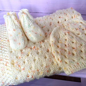 May include: A knitted baby set in a pale yellow colour with specks of orange, green, and yellow. The set includes a blanket, a hat, and booties.
