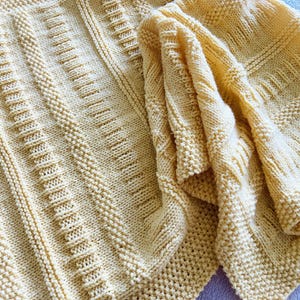 May include: A yellow knitted blanket with a cable knit pattern. The blanket is folded in half and has a soft, textured surface.