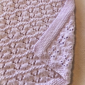 May include: A lavender knitted baby blanket with a delicate openwork pattern. The blanket is folded in half, showing the edge detail.