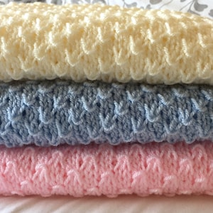 May include: Three crocheted baby blankets stacked on top of each other. The top blanket is cream, the middle blanket is light blue, and the bottom blanket is pink.