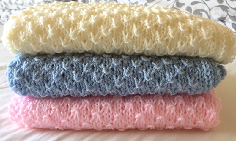 May include: Three crocheted baby blankets stacked on top of each other. The top blanket is cream, the middle blanket is light blue, and the bottom blanket is pink.