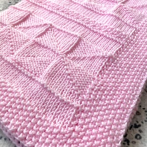 May include: A soft, pink knitted blanket with a textured, geometric pattern. The blanket is folded, showcasing the detailed stitch work and the delicate color. The overall design is a cozy, handmade aesthetic.