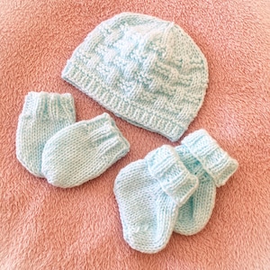 Op de afbeelding: Een lichtblauwe gebreide baby set, bestaande uit een muts en twee paar wanten. De set is gemaakt van zacht garen en wordt gepresenteerd op een roze ondergrond. De muts heeft een textuurpatroon.