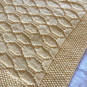 Könnte beinhalten: Eine gelbe gestrickte Babydecke mit einem Rautenmuster und einem einfarbigen Rand.