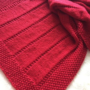 May include: A vibrant red hand-knitted blanket featuring a combination of textured garter stitch borders and vertical panels of stockinette stitch separated by delicate eyelet rows. The fabric displays a consistent deep crimson yarn throughout.