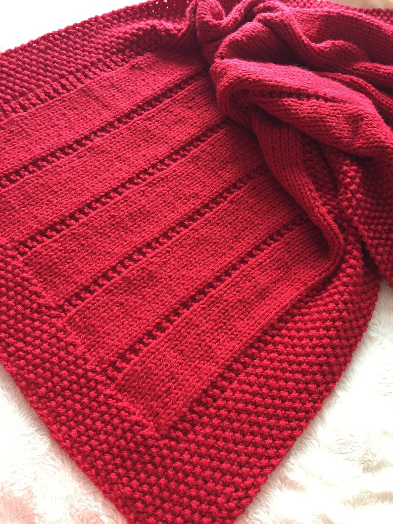 May include: A vibrant red hand-knitted blanket featuring a combination of textured garter stitch borders and vertical panels of stockinette stitch separated by delicate eyelet rows. The fabric displays a consistent deep crimson yarn throughout.