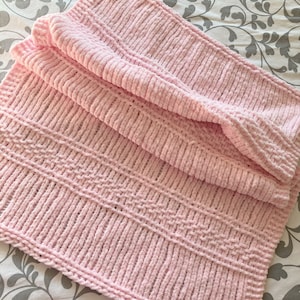 May include: A soft, pink, knitted blanket with a ribbed texture and a decorative patterned section. The blanket is folded, showcasing its plush, cozy appearance. The blanket is made of a thick, fuzzy material.