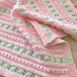 May include: A knitted baby blanket in pink and pale green. The blanket has alternating stripes of solid pink and pale green with a detailed openwork design. The blanket is folded, displaying the texture and intricate knitwork.