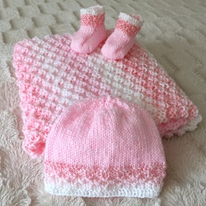 Könnte beinhalten: Eine gestrickte Babydecke, Mütze und Booties in Rosa und Weiß. Die Decke hat ein strukturiertes Stichmuster und die Mütze und Booties haben einen weißen Besatz.