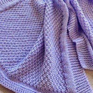 Może przedstawiać: Miękki, dziergany koc w jasnym kolorze lawendy. Koc ma wzorzysty ścieg i jest złożony, ukazując szczegółowe rzemiosło. Całkowity wygląd jest przytulny i zachęcający, odpowiedni do wystroju wnętrz.