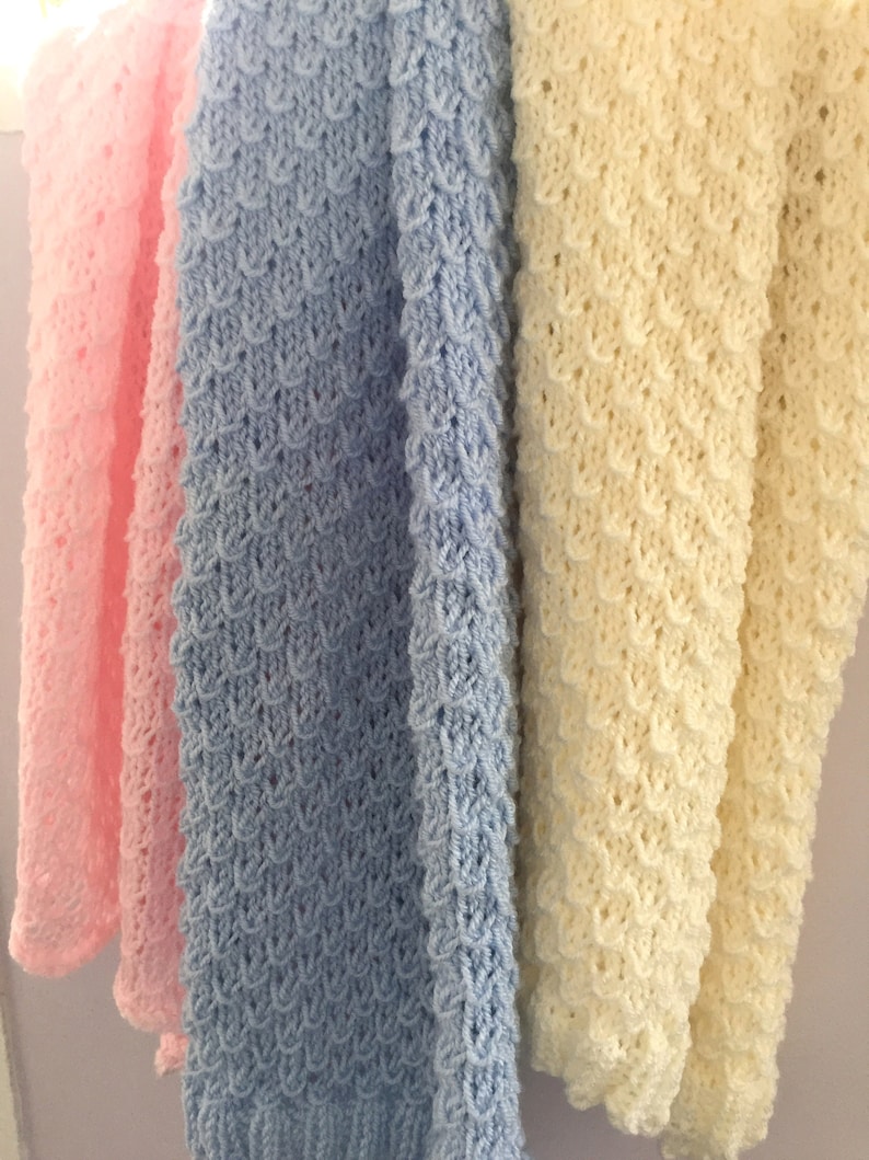 May include: Three crocheted baby blankets in pink, light blue, and cream colors. The blankets are made with a textured stitch and have a scalloped edge.
