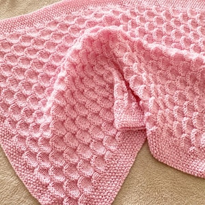 Peut inclure: Une couverture tricotée rose clair présentant un motif répétitif texturé en nid d'abeille ou en écailles sur toute la surface centrale, bordée par un point mousse simple. Le fil doux crée un effet tridimensionnel en relief sur le tissu.