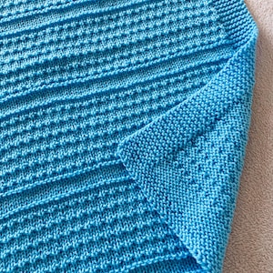 Puede incluir: Primer plano de una manta de punto en un vibrante azul turquesa. La manta presenta un patrón texturizado con secciones acanaladas horizontales y una esquina doblada, mostrando el detallado trabajo de puntada. La textura suave y acogedora es visible.