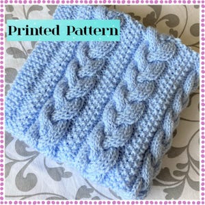 May include: A light blue knitted blanket with a cable knit pattern. The blanket has a textured surface and the words "Printed Pattern" are displayed in a rectangular box at the top. The blanket is folded and appears soft and cosy.
