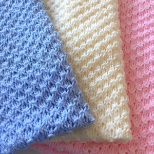 May include: Three crocheted baby blankets in blue, white, and pink. The blankets are made with a textured stitch and are folded on top of each other.