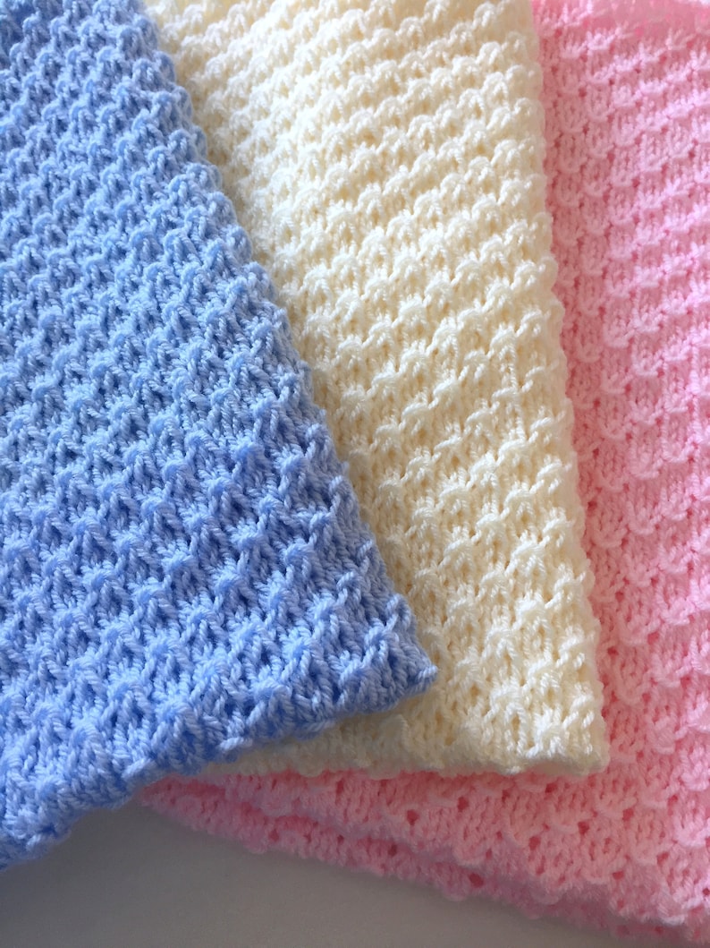 May include: Three crocheted baby blankets in blue, white, and pink. The blankets are made with a textured stitch and are folded on top of each other.