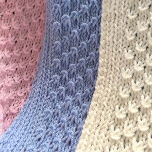 May include: Close-up of three knitted fabric swatches in pink, light blue, and white. The swatches have a textured, lacy pattern.