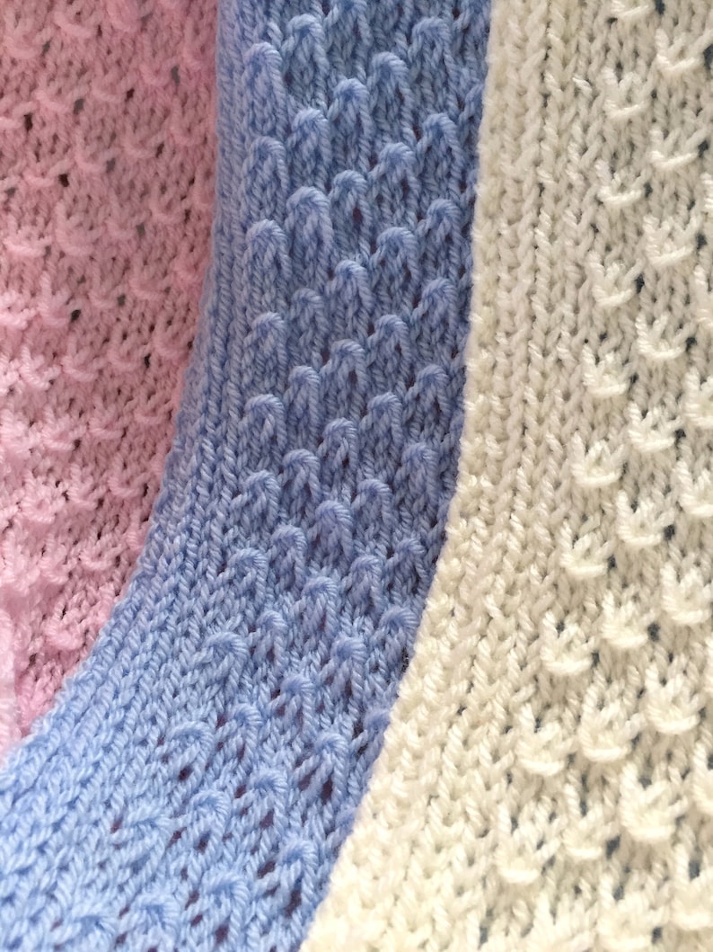May include: Close-up of three knitted fabric swatches in pink, light blue, and white. The swatches have a textured, lacy pattern.