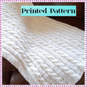 May include: A cream-coloured knitted blanket featuring a cable knit design. The blanket is draped over a brown surface. The text "Printed Pattern" is displayed on a light blue rectangle.