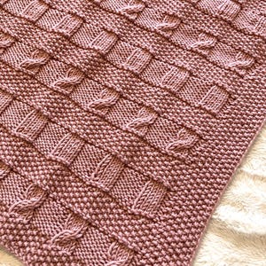 May include: A soft, pink knitted blanket with a cable knit pattern. The blanket is made with a fine yarn and has a delicate texture.