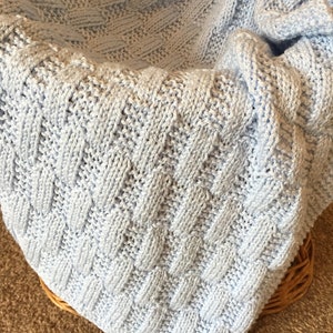 May include: A light blue knitted baby blanket with a woven pattern. The blanket is folded and draped over a wicker basket.