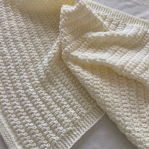May include: A white knitted blanket with a textured pattern. The blanket is folded in half and is lying on a white surface.