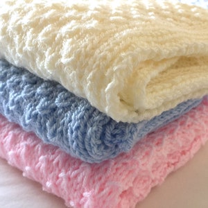 May include: Three knitted baby blankets stacked on top of each other. The top blanket is white, the middle blanket is light blue, and the bottom blanket is pink.