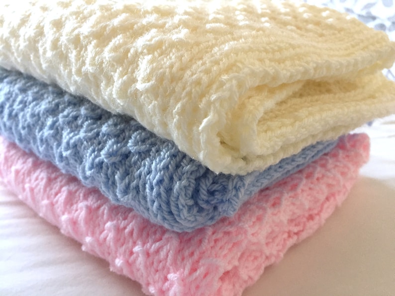 May include: Three knitted baby blankets stacked on top of each other. The top blanket is white, the middle blanket is light blue, and the bottom blanket is pink.