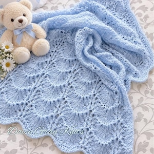 May include: A light blue knitted blanket featuring a scalloped lace pattern with openwork details. A beige plush teddy bear with a light blue ribbon around its neck and a small cluster of white daisy flowers are positioned on the blanket.