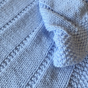 May include: A light blue knitted blanket featuring a textured pattern of vertical columns. The fabric displays a combination of smooth stockinette stitches and raised, bumpy stitch sections that create a repeating striped design throughout.