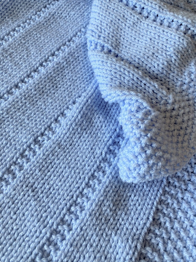 May include: A light blue knitted blanket featuring a textured pattern of vertical columns. The fabric displays a combination of smooth stockinette stitches and raised, bumpy stitch sections that create a repeating striped design throughout.