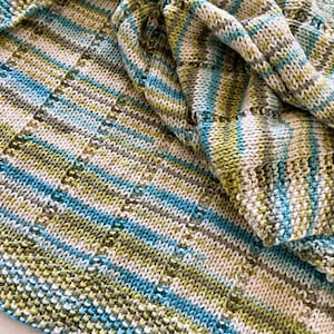 May include: A hand-knitted blanket featuring a variegated yarn pattern in shades of lime green, turquoise blue, white, and soft grey. The textile displays a consistent stockinette stitch with vertical columns of eyelet lace detailing throughout the fabric.