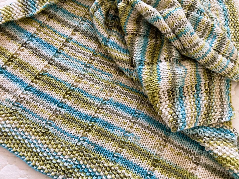 May include: A hand-knitted blanket featuring a variegated yarn pattern in shades of lime green, turquoise blue, white, and soft grey. The textile displays a consistent stockinette stitch with vertical columns of eyelet lace detailing throughout the fabric.