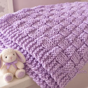May include: A light purple knitted blanket featuring a textured basketweave pattern. Beside the blanket sits a small, cream-colored plush rabbit toy with long ears, wearing a light purple ribbon around its neck. The text Daisy Gray Knits is visible.