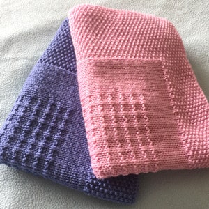 Puede incluir: Dos mantas de bebé tejidas, una de color morado claro y otra de color rosa claro. Ambas mantas tienen un patrón de punto texturizado.