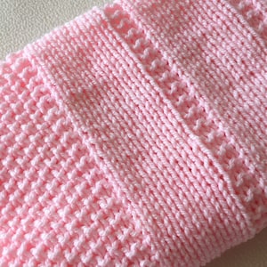 May include: A soft, light pink knitted blanket featuring a textured pattern with alternating vertical sections of a basketweave stitch and a delicate, openwork lace stitch, creating a uniform and repeating design across the entire fabric surface.