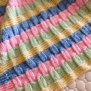 May include: A hand-knitted blanket featuring a textured basketweave pattern in pastel colors including light blue, soft pink, pale yellow, and mint green. The yarn has a speckled appearance with tiny flecks of vibrant colors throughout the fabric.