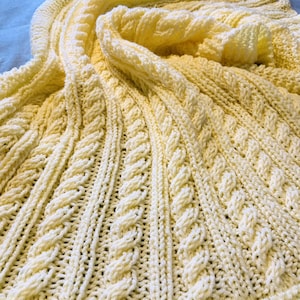 Op de afbeelding: Een zacht, geel gebreid babydekentje met een kabelbreipatroon.