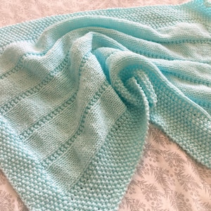 May include: A light blue knitted blanket featuring a textured garter stitch border and a central panel with alternating vertical columns of stockinette stitch and horizontal rows of eyelet detailing, creating a soft and uniform appearance.