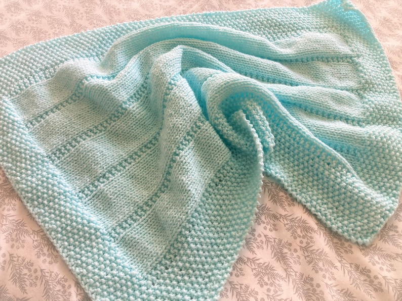May include: A light blue knitted blanket featuring a textured garter stitch border and a central panel with alternating vertical columns of stockinette stitch and horizontal rows of eyelet detailing, creating a soft and uniform appearance.