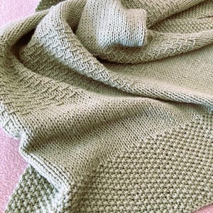 May include: A sage green knitted shawl with various textured patterns. The shawl is draped, showcasing the different stitch designs and the soft, flowing fabric. The color is a muted, earthy tone, suitable for a variety of outfits.