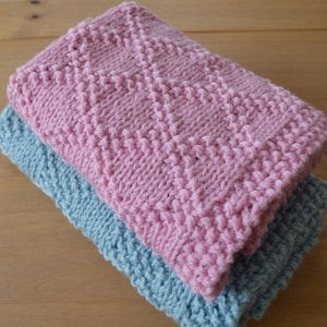May include: Two knitted dishcloths, one pink and one blue, with a textured diamond pattern. The cloths are stacked on top of each other.