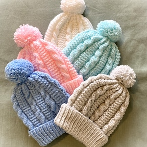 以下が含まれることがあります： 白、ピンク、水色、ベージュなど、さまざまな色のポンポンが付いたニットのベビーハットが5つあります。