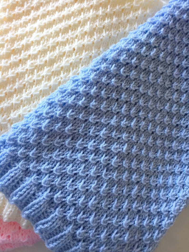 May include: Two knitted baby blankets, one in light blue and one in cream. The blankets are made with a textured stitch.